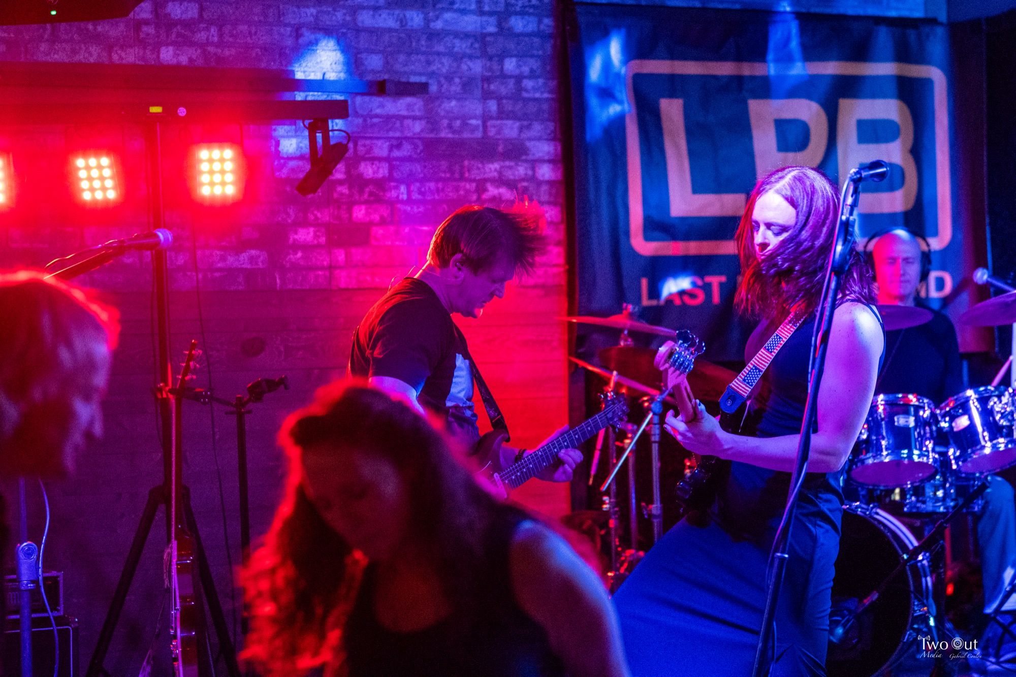 Guitarist mid head-bang next to the bassist at Buzzed Crow, audience heads visible in the foreground