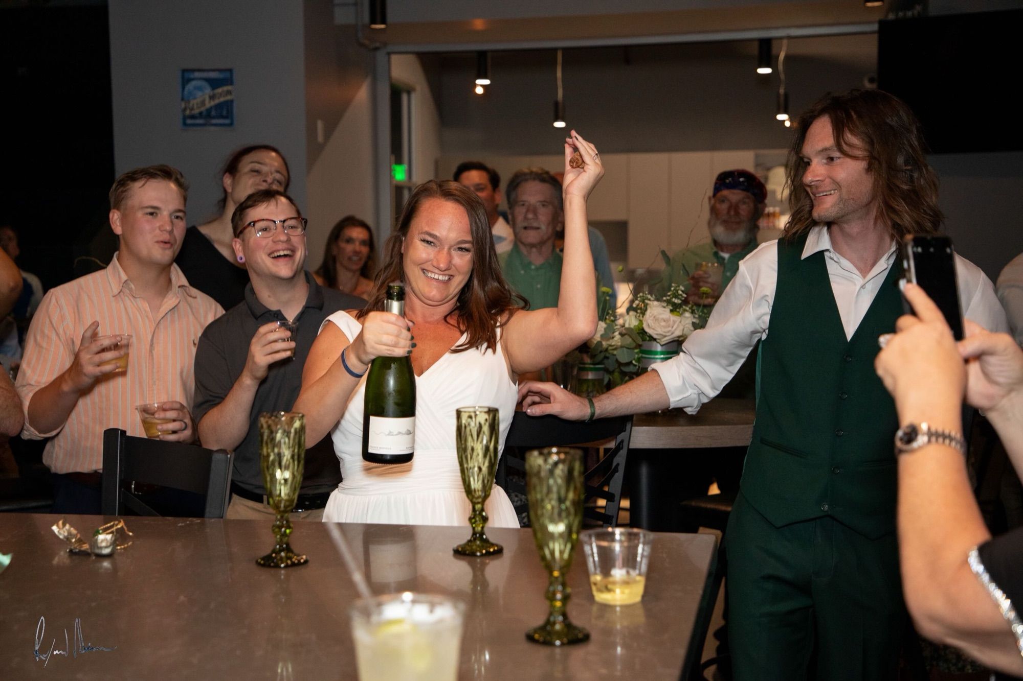 Chelsey popping a bottle of champagne as guests cheer at the reception