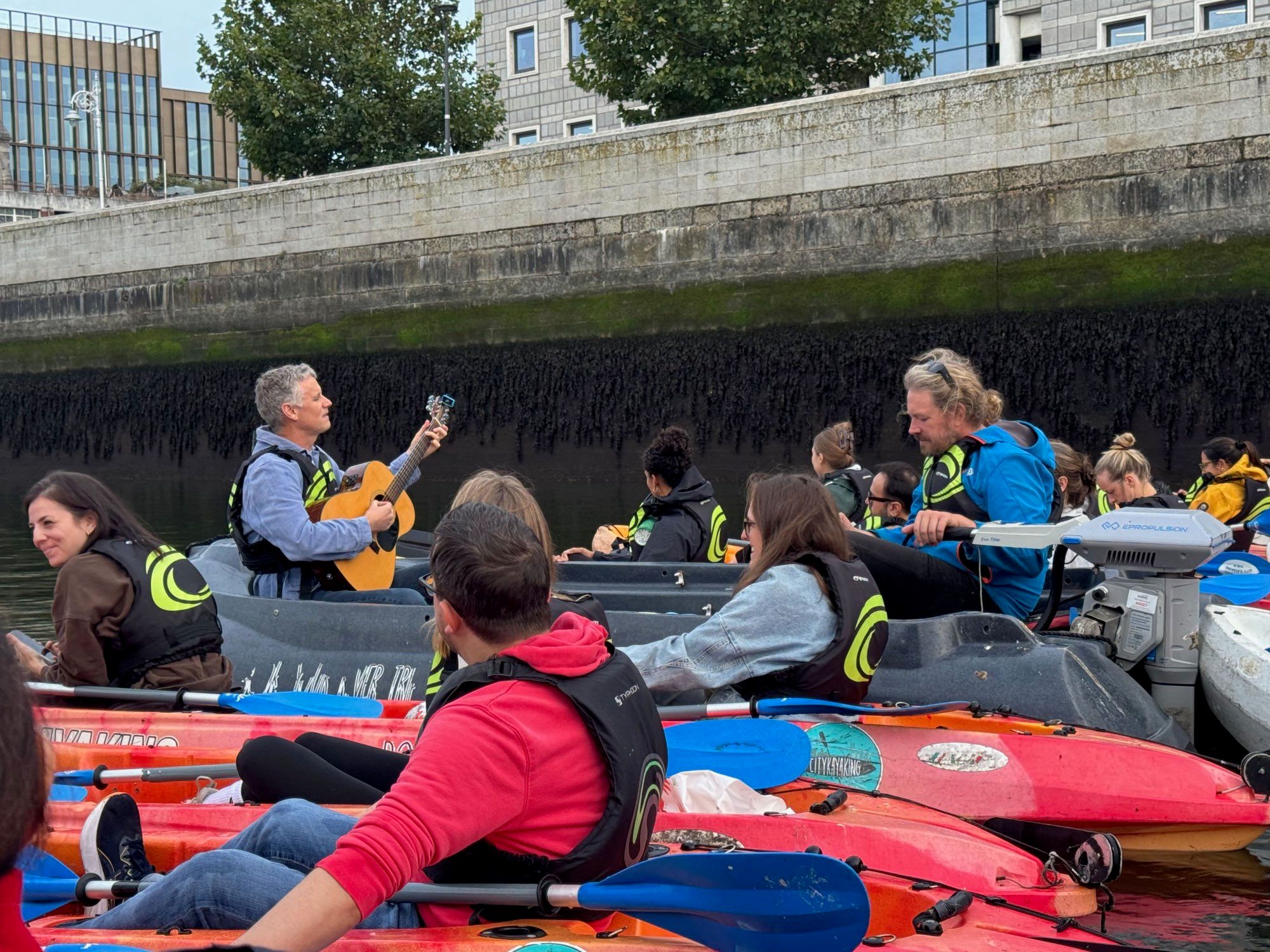 A musician playing guitar surrounded by kayakers in branded life jackets