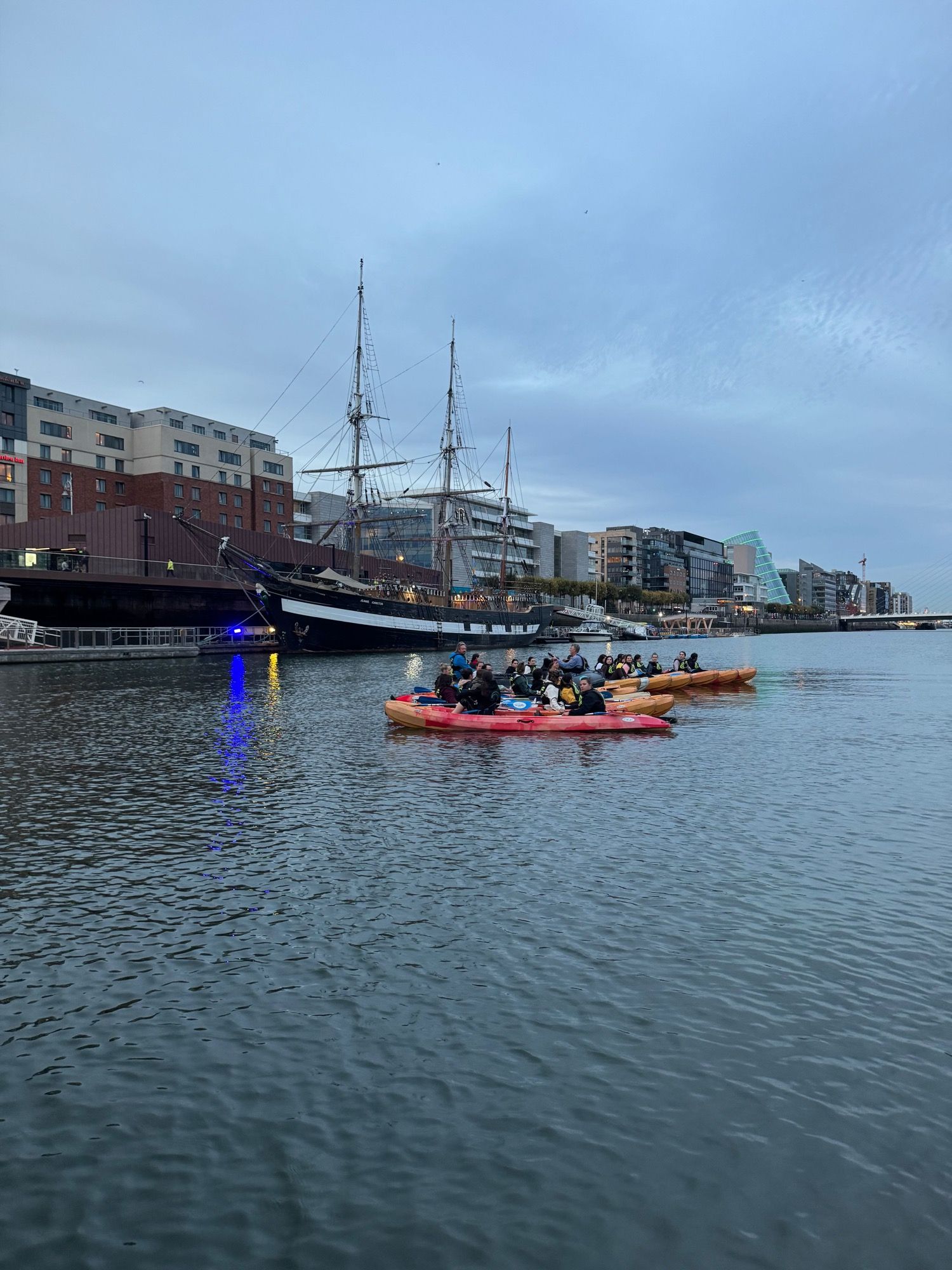 Kayakers paddling past a tall ship moored in Dublin's docklands at dusk